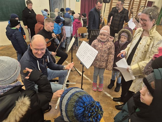 Bild: Viele Kinder umringen einen Musiker, der ihnen ein Musikinstrument zeigt. 